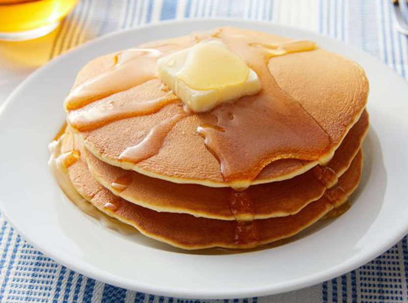 Stack of fluffy Cracker Barrel pancakes topped with butter and syrup on a white plate with blue checkered tablecloth.